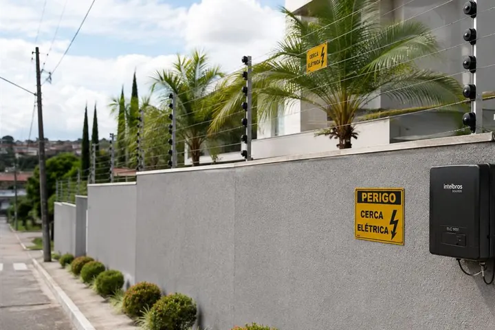 Cerca elétrica no alto de um muro residencial, vista da calçada, com a placa de perigo na parede