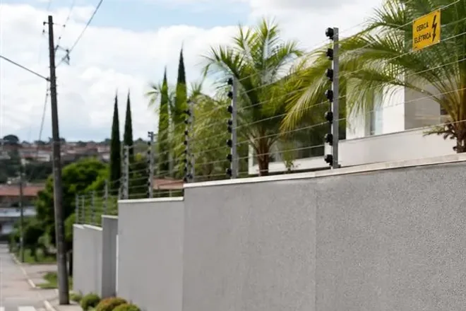 Muro alto de casa visto da rua, com hastes, isoladores e fios da cerca elétrica no topo e placa de aviso